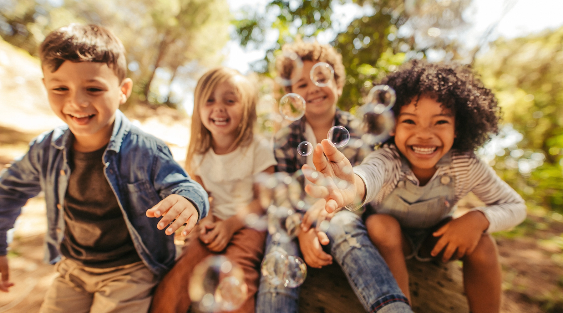 A joyful outdoor scene of four children playing with bubbles, smiling and leaning toward the camera in a sunlit natural setting.